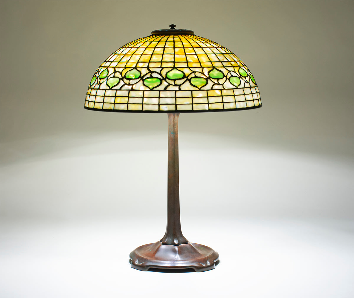 a geometric tiffany studios lamp, leaded glass shade in the &quot;acorn&quot; pattern, the background a pale creamy yellow grid of glass with a horizontal decorative band of stylized leaves alternating pointing up and down in green glass