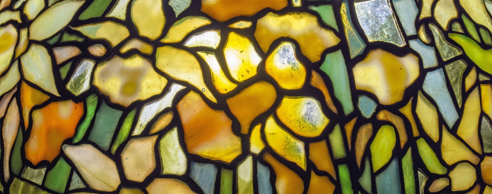 A detail shot of a Tiffany Studios Daffodil Lamp, showing the leaded glass shade with mottled light yellow daffodils against a background of light green leaves and pale blue sky.