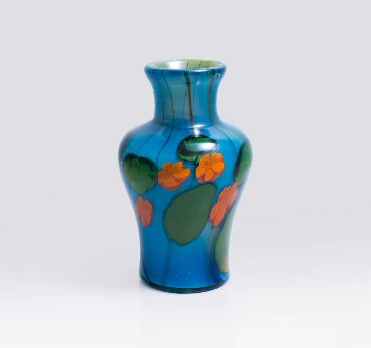 a favrile glass paperweight vase by tiffany studios, blue background with vibrant red-orange nasturtium flowers surrounded by large round green leaves swirl above a background of vibrant blue glass. the vase is of baluster form.