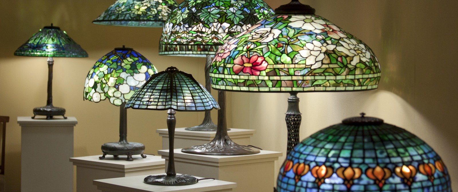 A photograph showing a grouping of Tiffany Lamps in a Lillian Nassau LLC exhibition, each with a leaded glass shade in vibrant colors depicting various flowers and geometric patterns.