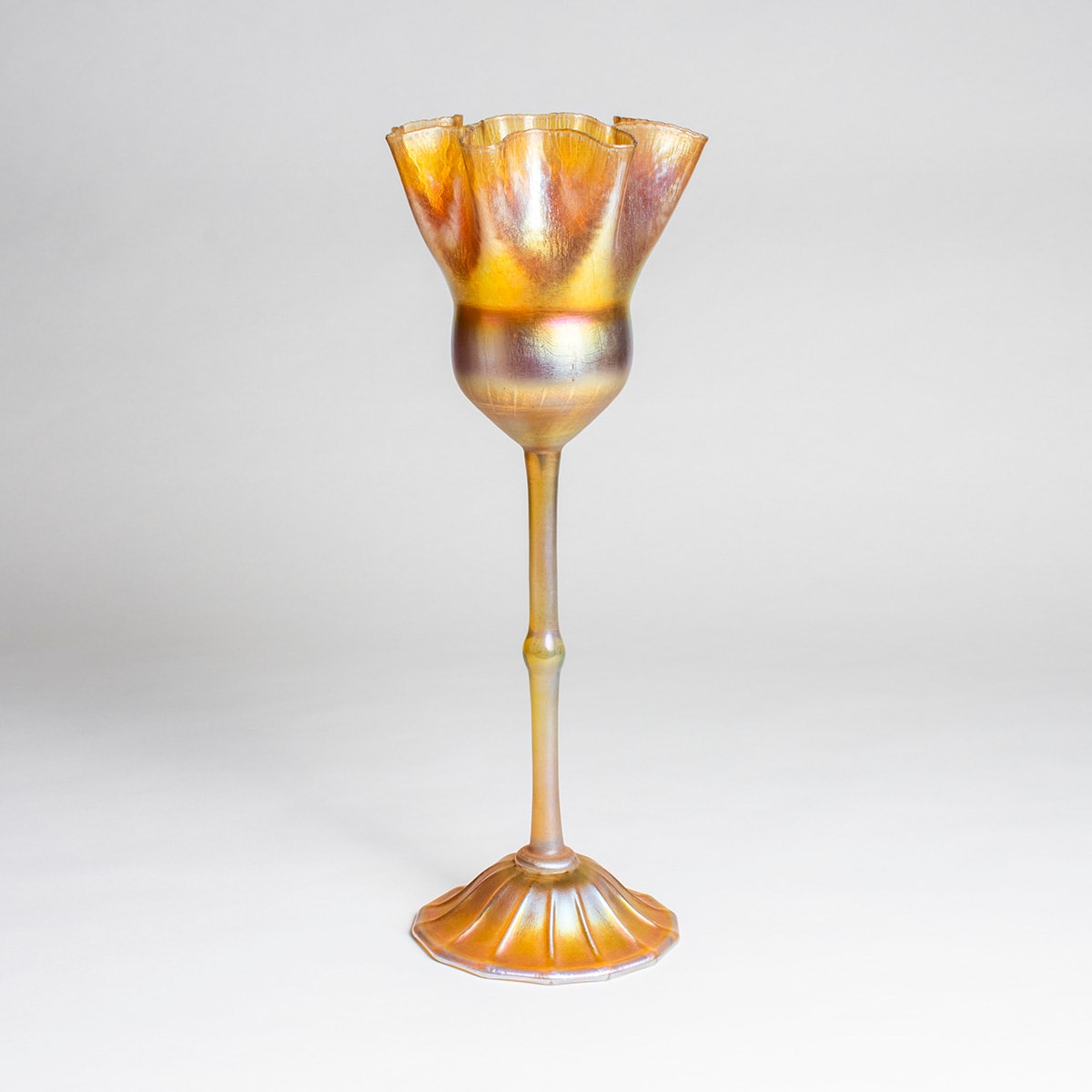 a flower form in gold iridescent favrile glass, made by tiffany studios around 1904, on a domed foot, the thin stem with a knob in the middle, the flower cup rounded at the base with pinched ruffles