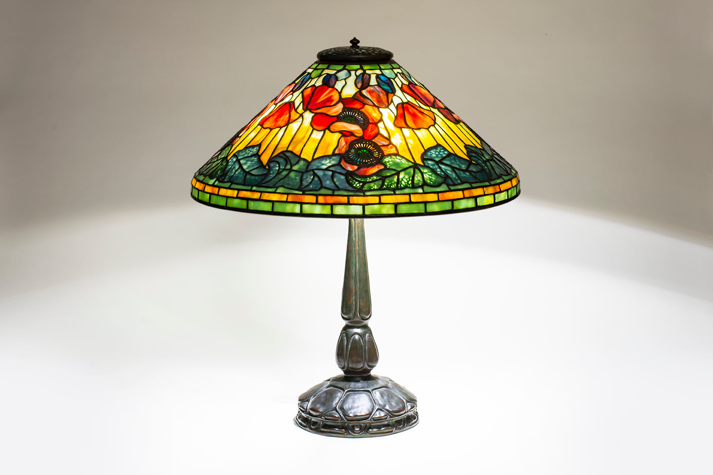 a leaded glass Tiffany Lamp depicting red poppies with pierced metal filigree mounted on top of the glass representing the centers of the flowers, against a background of variegated yellow glass, with poppy buds in blue-red glass near the top of the shade, the lower edge of the shade with green leaves, the vines formed by pierced metalwork mounted to the interior of the shade.