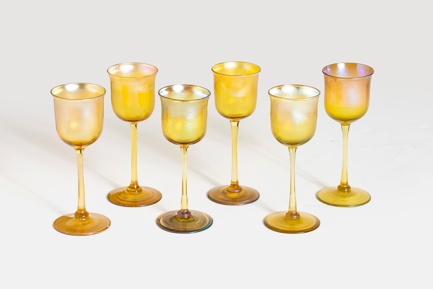 set of 6 gold iridescent tiffany favrile glass drinking glasses for liqueurs, yellowy base glass with bright shiny rainbow silver iridescence, the glasses with shallow slightly flared cups, thin stems and rounded flat feet.