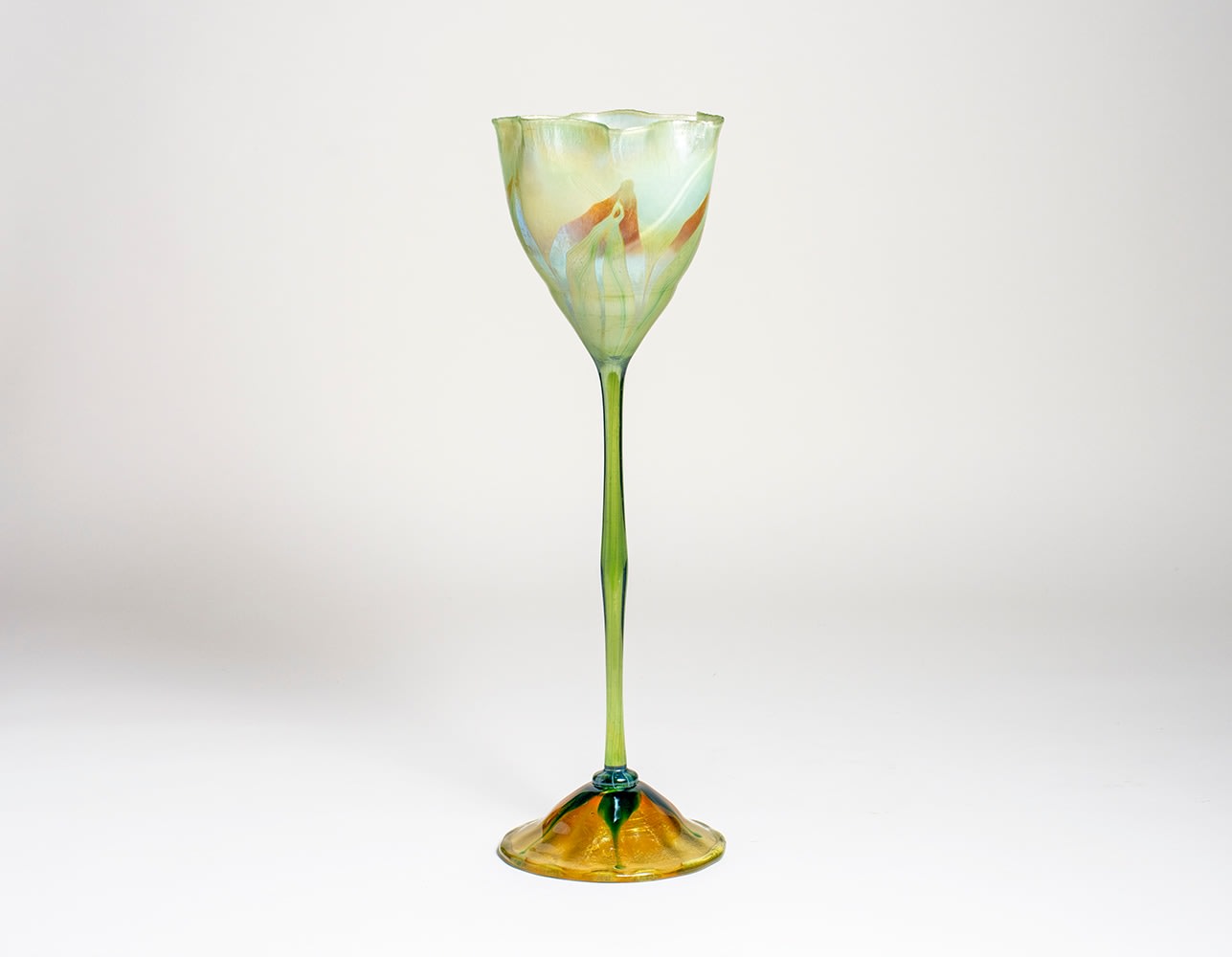 an early blown tiffany glass &quot;flower form&quot; vase, an art glass piece meant to be a piece of sclupture instead of a vase, the thin stem rising to a ruffled flower cup decorated with iridescent pulled leaf decoration, the base glass a pale opalescent green