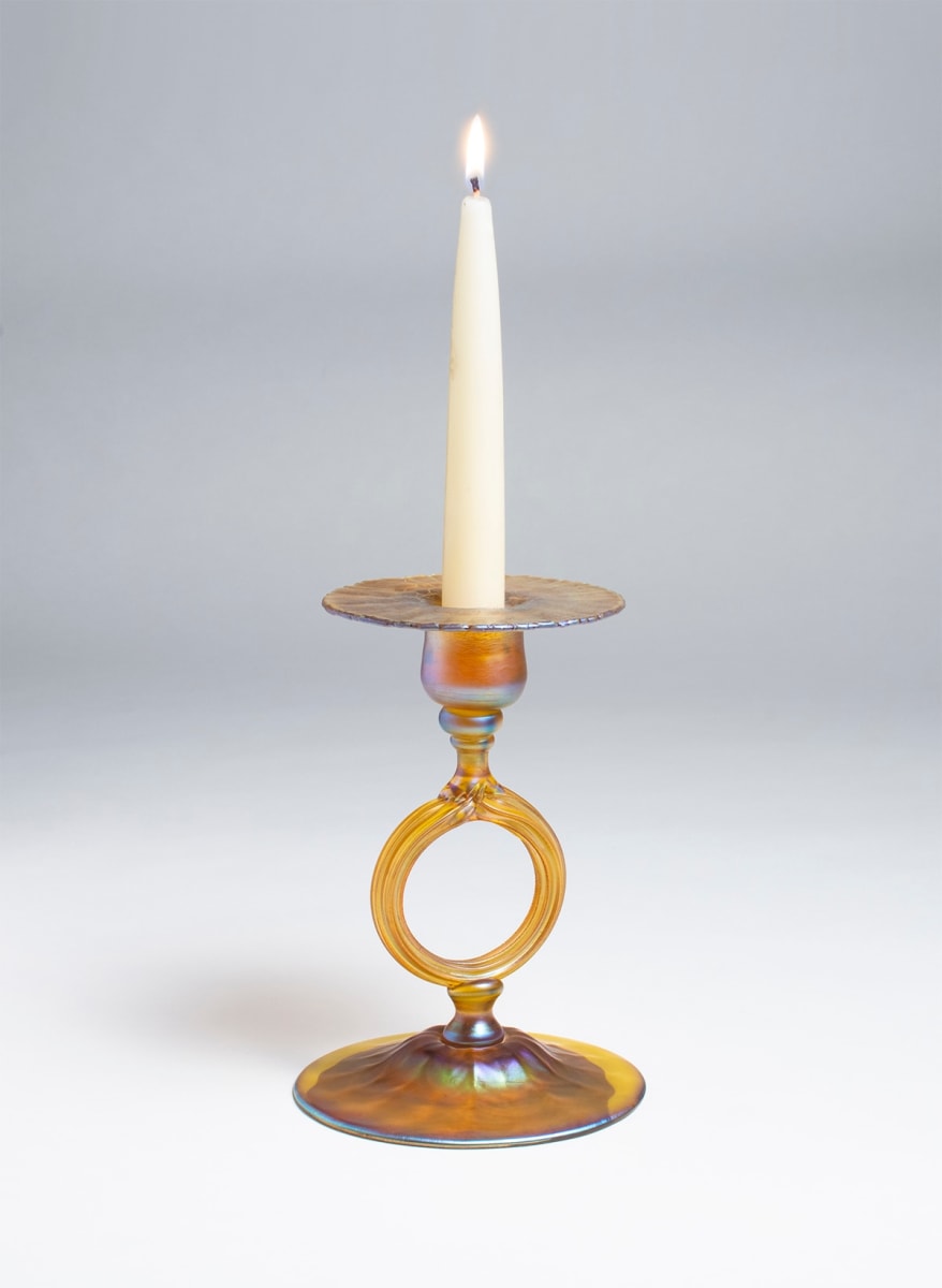 a gold iridescent Tiffany Favrile Glass Candle Stick, on a domed foot, with an open circular ring for the &quot;stem&quot; rising to support a flared candle cup - shown here with a lit ivory candle.