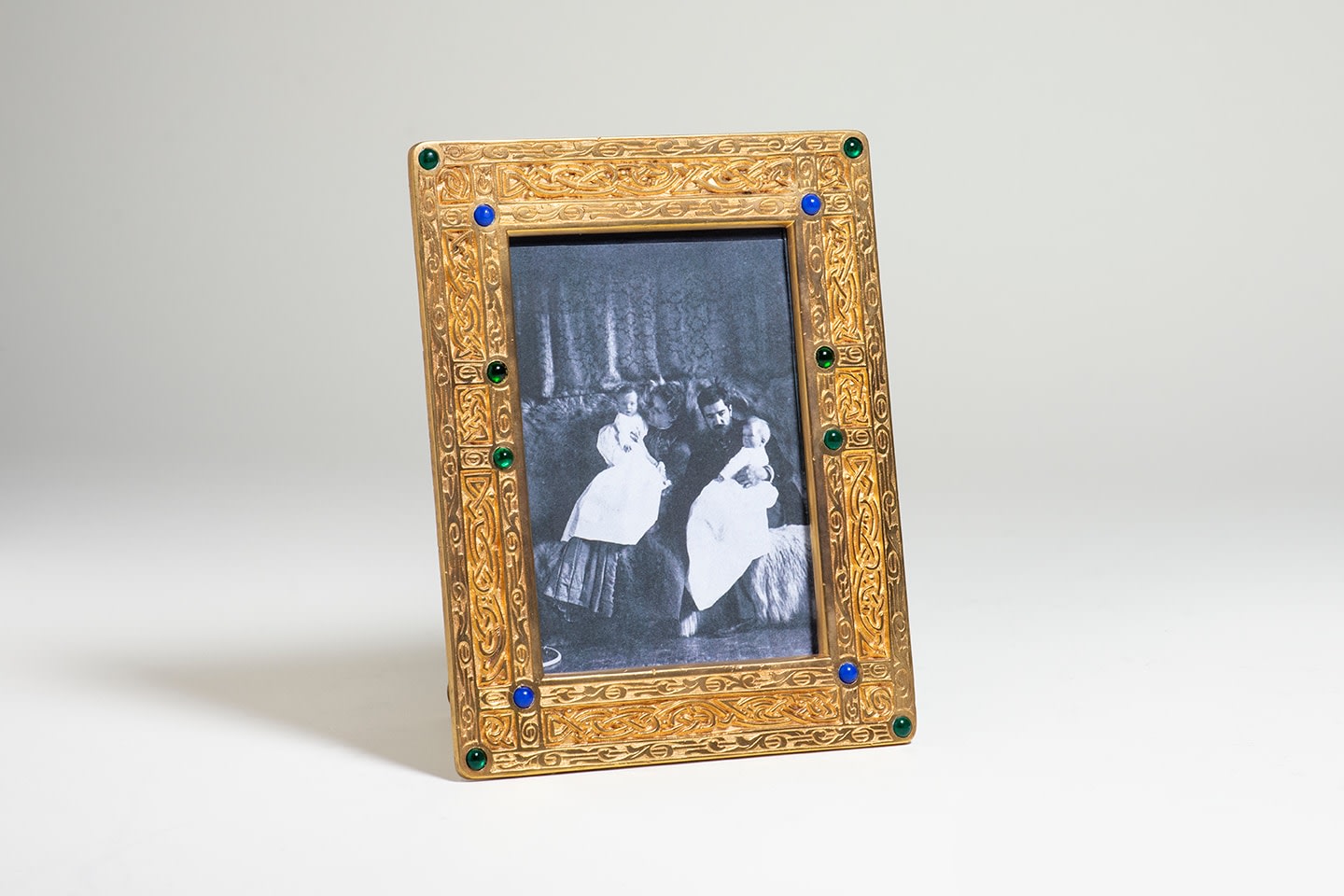 A vertically oriented rectangular picture frame in gilt bronze decorated on the borders with a deeply recessed intricate celtic inspired interlace or strapwork design, part of the Ninth Century desk set by Tiffany Studios, with inset circular pressed Tiffany Glass &quot;gems&quot; in green and blue glass inset at the corners and along the borders