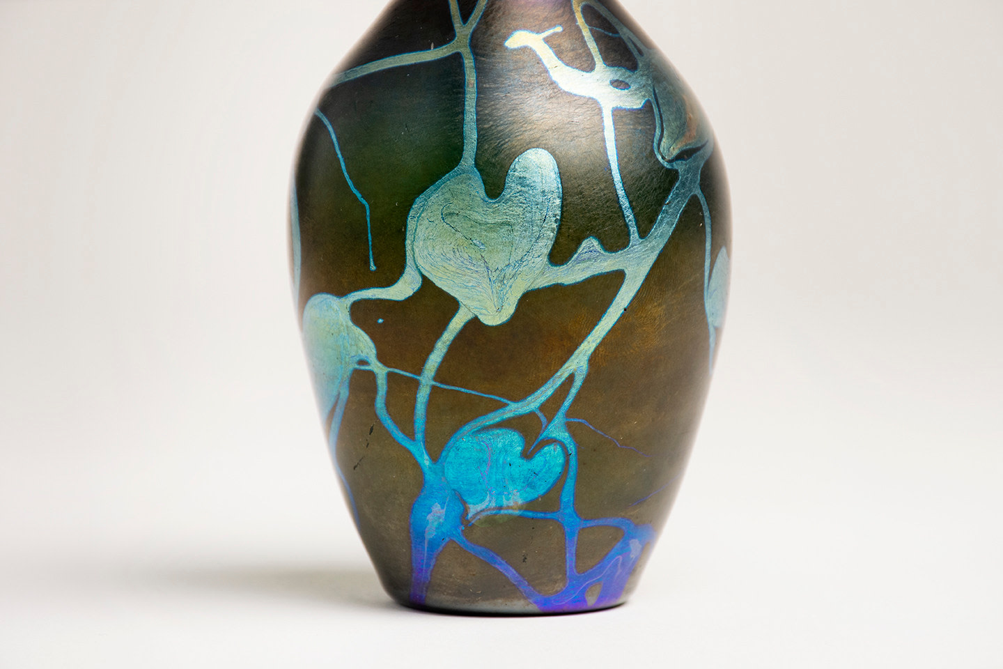 A detail shot of a blown Tiffany Favrile Glass cabinet vase, the vase body formed by deep almost black glass with soft sheen of iridescence, decorated with a leaf and vine motif in shinier iridescent silvery blue glass