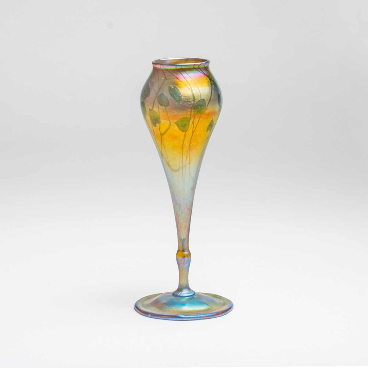 a gold iridescent tiffany glass flower form vase, the rounded flower cup decorated with a motif of green swirling thin vines and heart shaped leaves