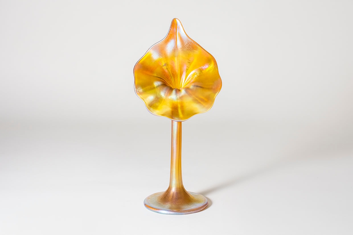 a gold iridescent tiffany glass flower form known as the Calla, the flattened circular foot rising to a thin cylindrical stem which spreads into a teardrop shaped flower face with ruffled edges and pointed tip, referencing the Calla Lily flower