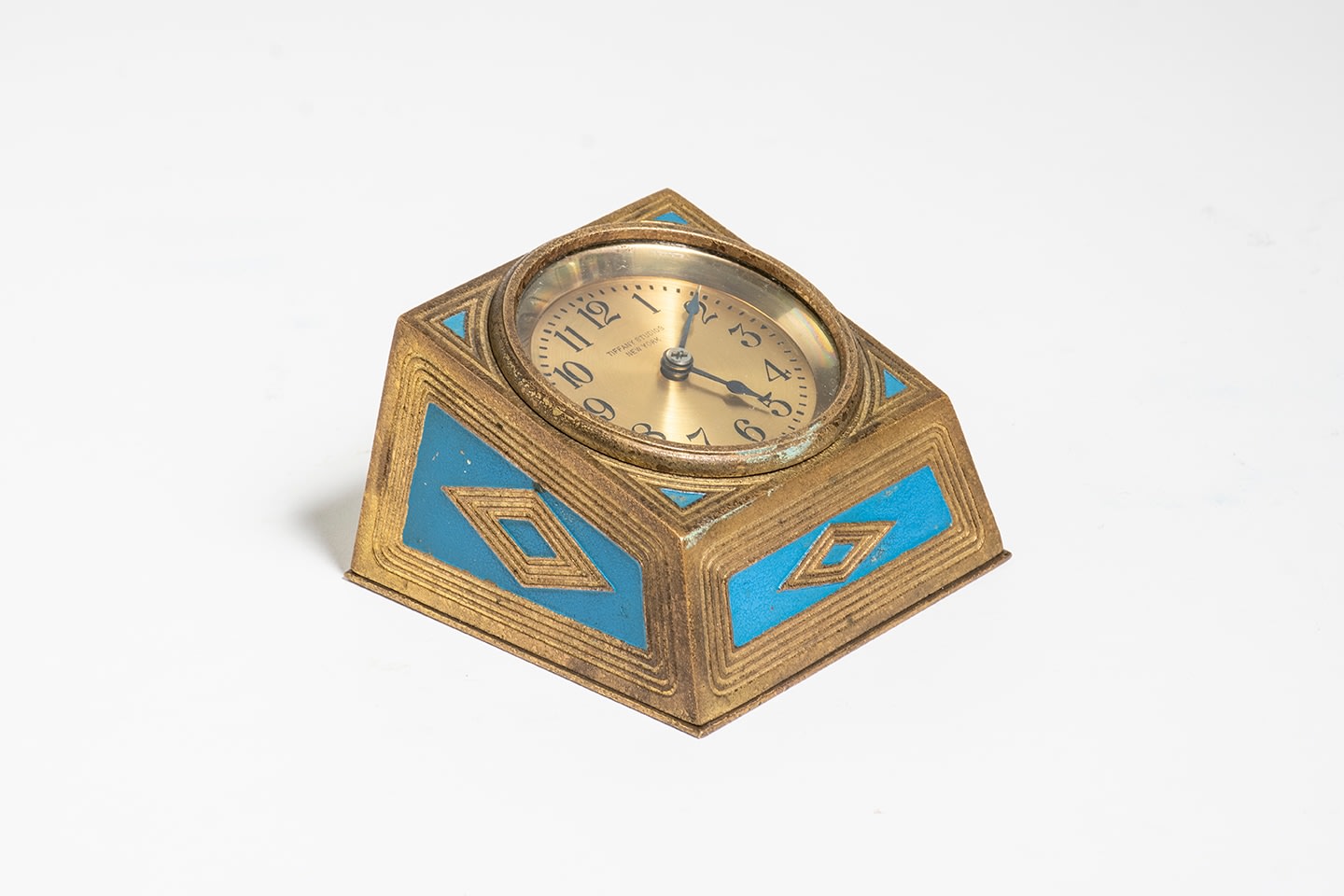 a low squared desk clock with round clock face marked TIFFANY STUDIOS NEW YORK, in gilt bronze, the four flattened sides with raised linear and diamond motifs inset with soft blue enamel