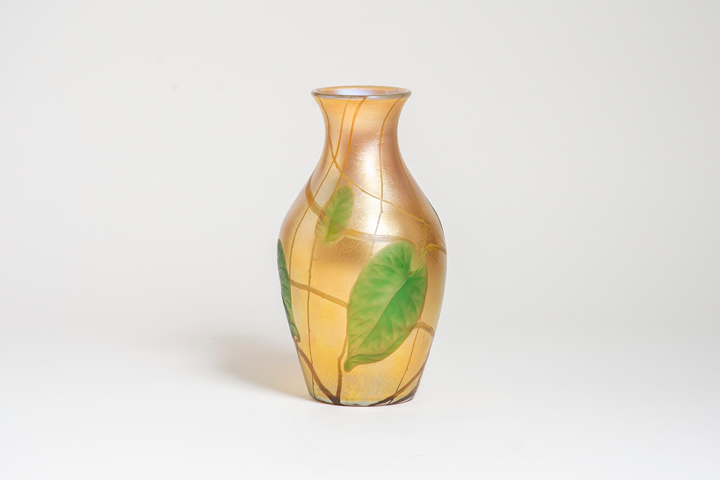 a baluster form vase in gold iridescent Tiffany Glass, decorated with a motif of swirling vines and heart-shaped leaves in green glass which have been wheel-carved to have additional decoration of veins and thorns