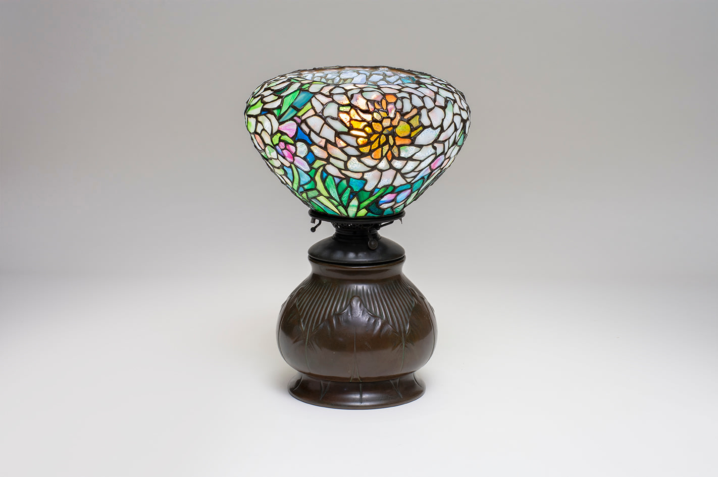 an early peony tiffany lamp, the shade a torchiere shape based on earlier blown glass shades, on a bulbous bronze base decorated with tobacco leaf motif originally intended as an oil lamp base