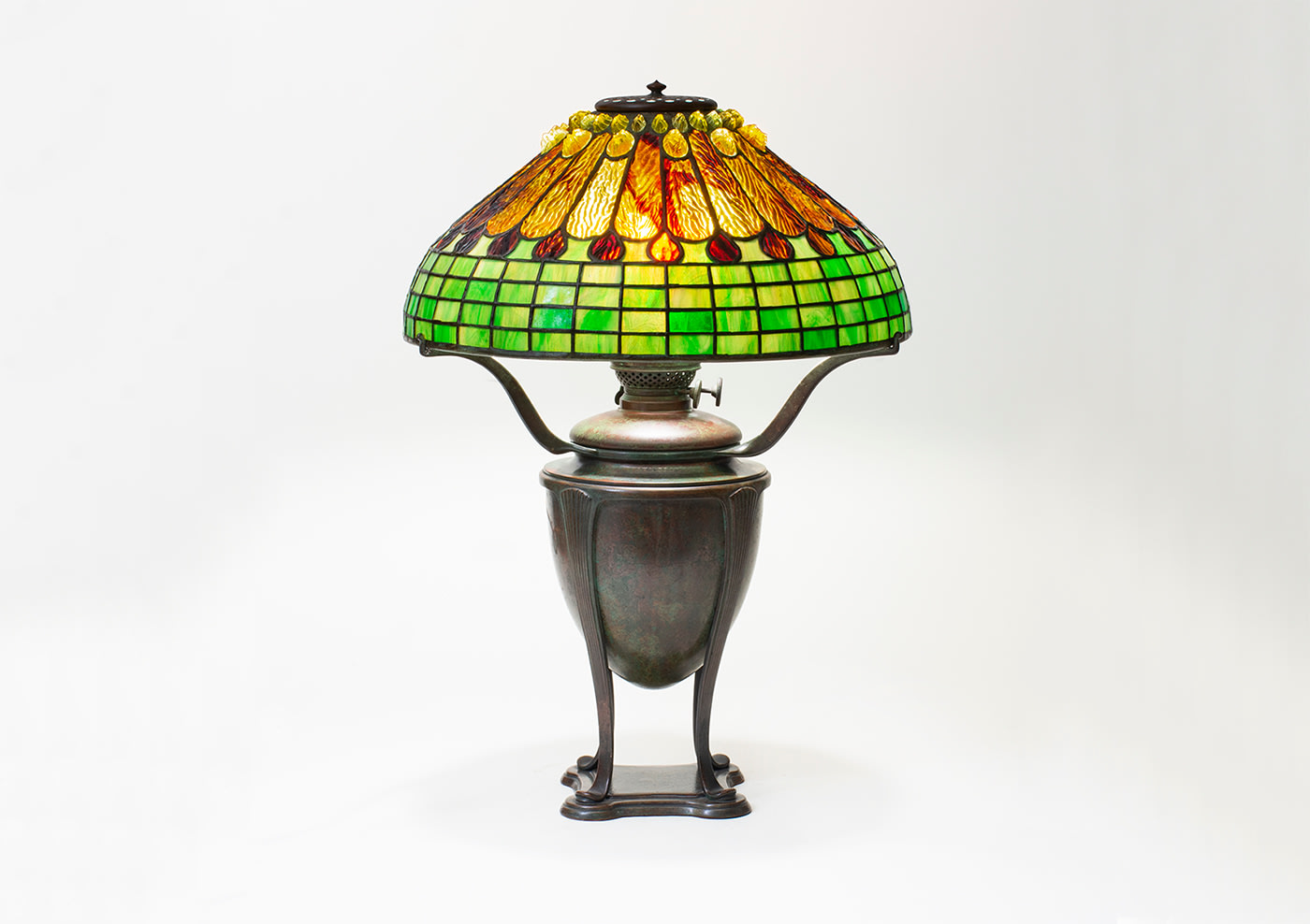 an early tiffany lamp with geometric shade formed by panels of &quot;rippled&quot; tiffany glass in shades of amber and deep orange-red with the ripples arranged so that the shade resembles the feathers of a bird, the crown of the shade with pressed tiffany glass jewel &quot;chunks&quot;, on a bronze urn shaped base in rich brown-green patina