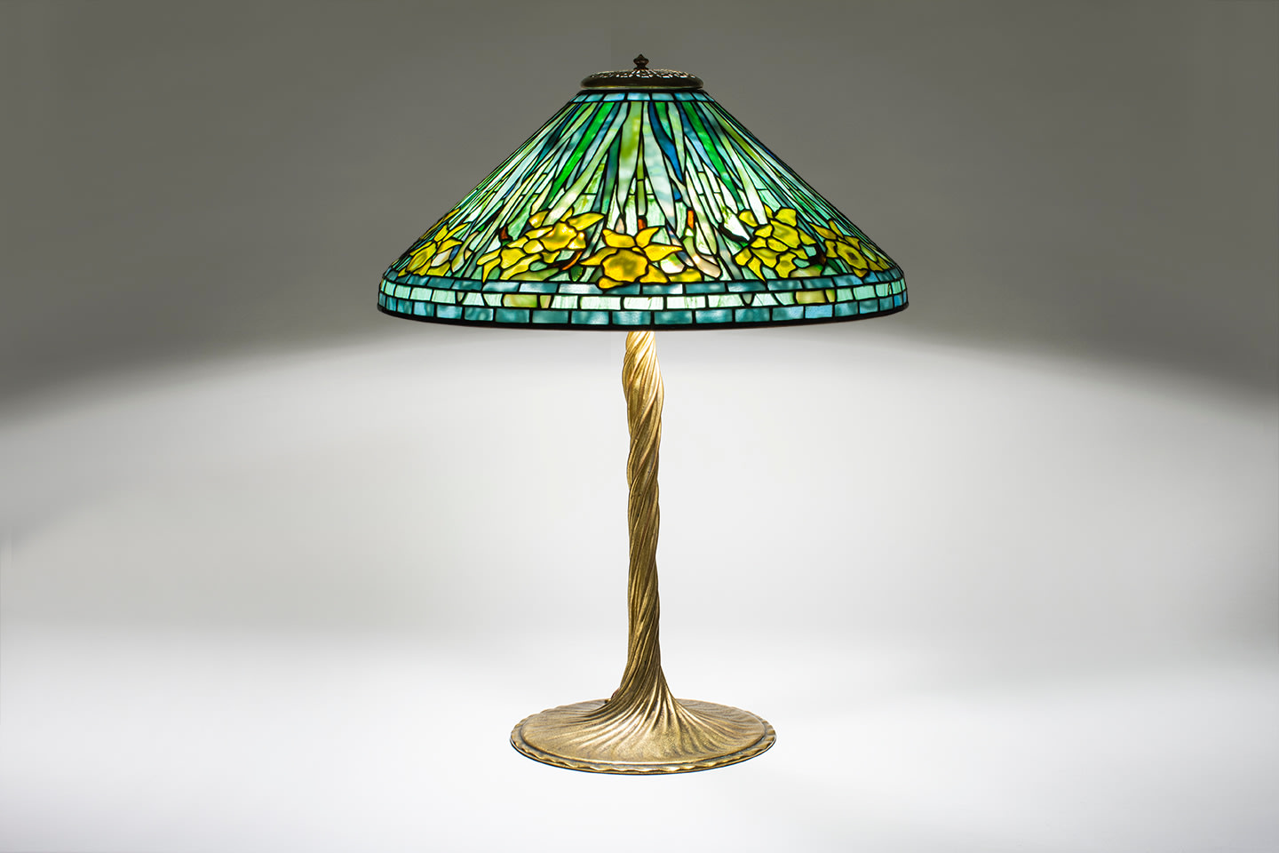 an original tiffany lamp with a leaded glass shade depicting yellow daffodils against spiky green leaves and a blue background, on a gilt bronze base which is a sculptural representation of a twisted vine