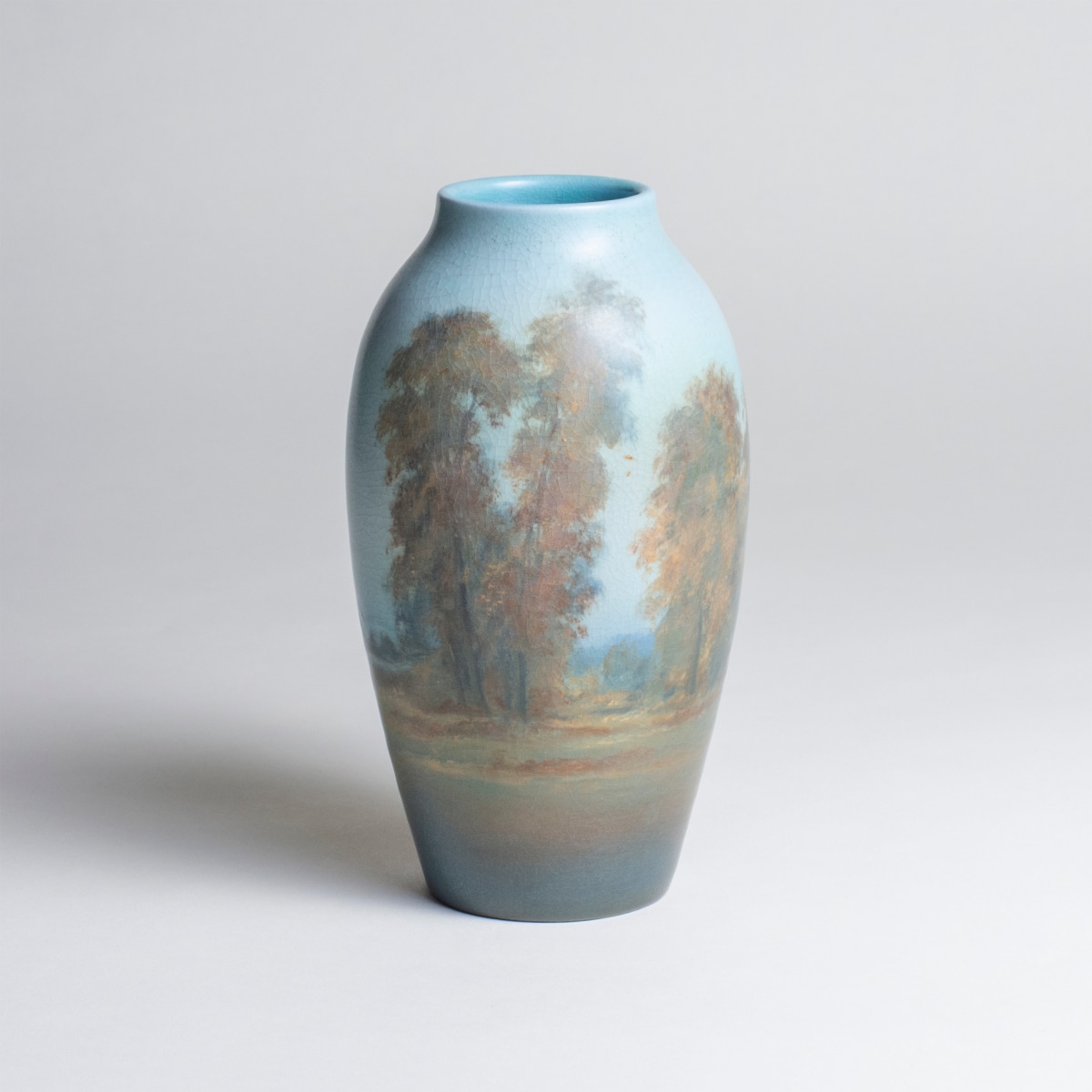 a landscape vase of the ohio river or american landscape by rookwood pottery artist ed diers, showing a fall scene with trees in autumn foliage with snow capped mountains in the distance, in the hazy scenic vellum glaze