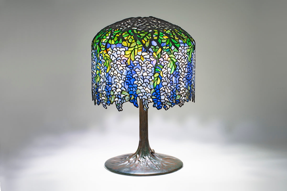 a leaded glass original tiffany lamp with shade depicting pendulous lavender and soft blue wisteria blossoms, with irregular lower edge, on a bronze &quot;tree trunk&quot; base