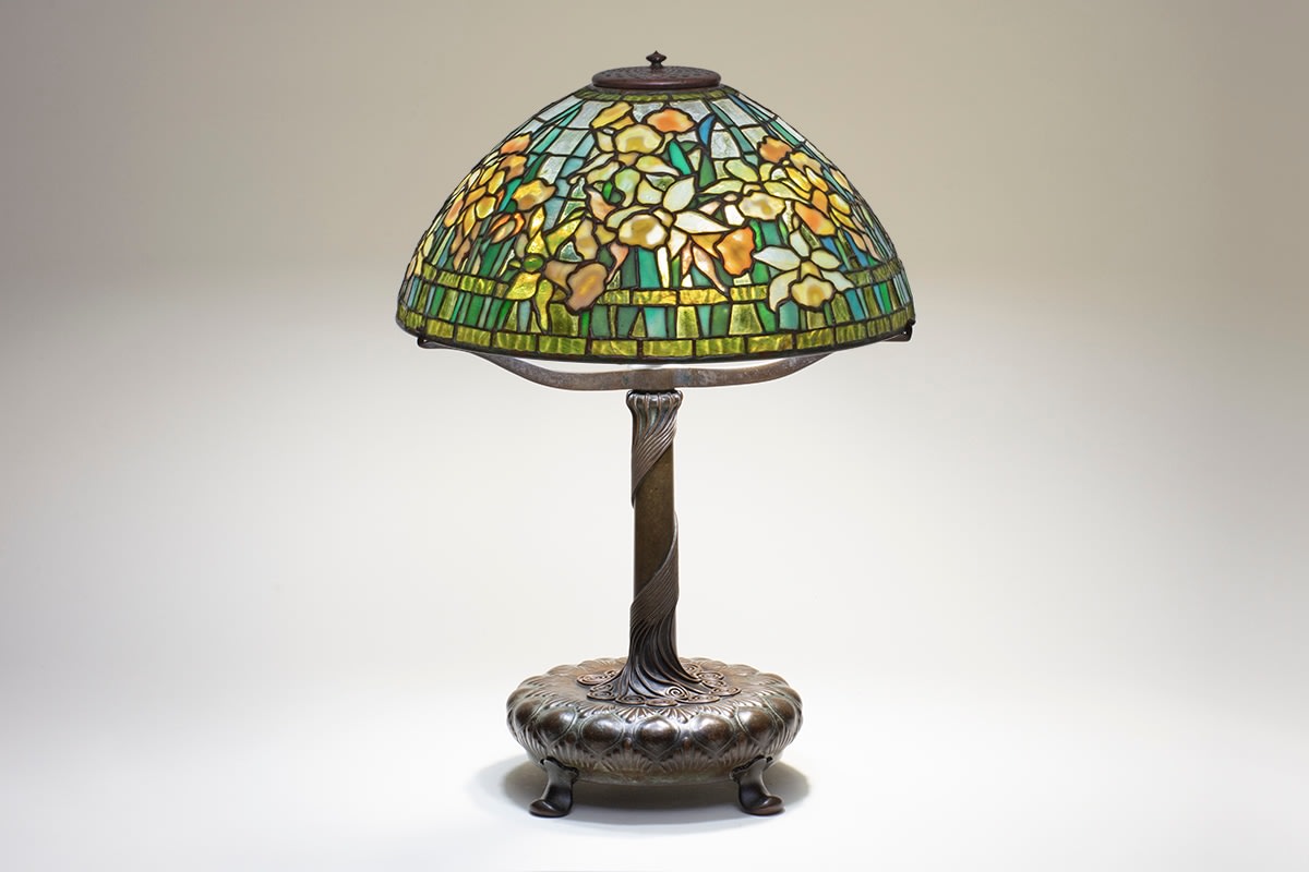 an authentic tiffany lamp with a domed leaded glass shade depicting yellow daffodils with green spiky leaves, against a transparent clear glass &quot;sky&quot;