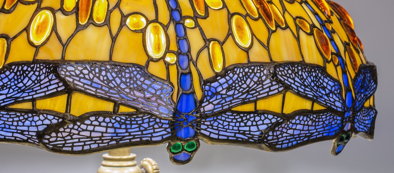 A detail shot of an original leaded glass Tiffany Lamp shade depicting blue dragonflies with rounded teal eyes, against a background of streaky golden yellow glass with rounded yellow pressed glass jewels