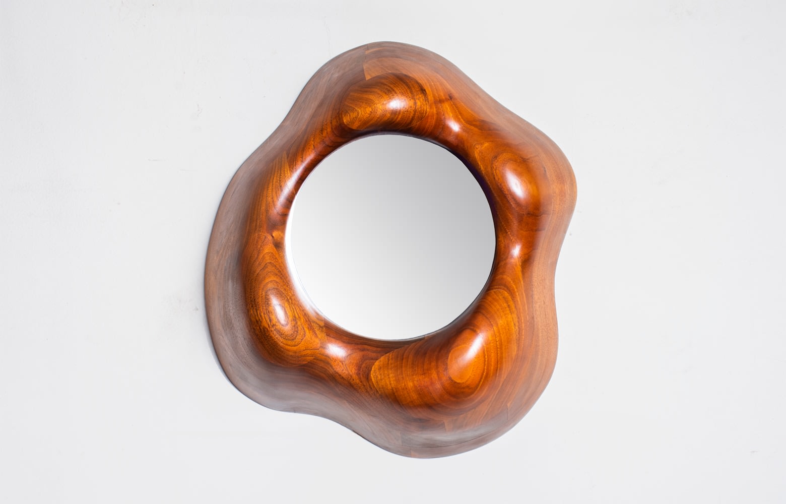 a stacked and laminated carved wooden mirror of rounded biomorphic form, in the style of his 1960s-70s works, by american master craftsman wendell castle, with an inset circular mirror, intended to hang from a wall.