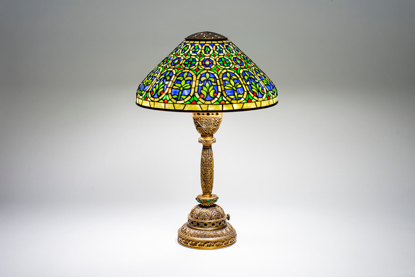 an original tiffany lamp from the venetian desk set, the complex gilt bronze base matching the desk set's intricate openwork pattern of scrollwork and motif of ermines, with inset green rounded glass jewels, paired with a conical shaded of leaded tiffany glass depicting alternating medallions of palmettes and fleur de lys with a geometric motif in blue, red, and green glass