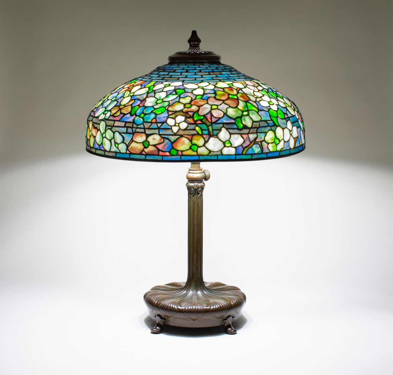 a leaded glass tiffany lamp, the shade depicting dogwood blossoms in a variety of pastel colors, produced by tiffany studios, on a bronze decorated cushion table base in brown patina