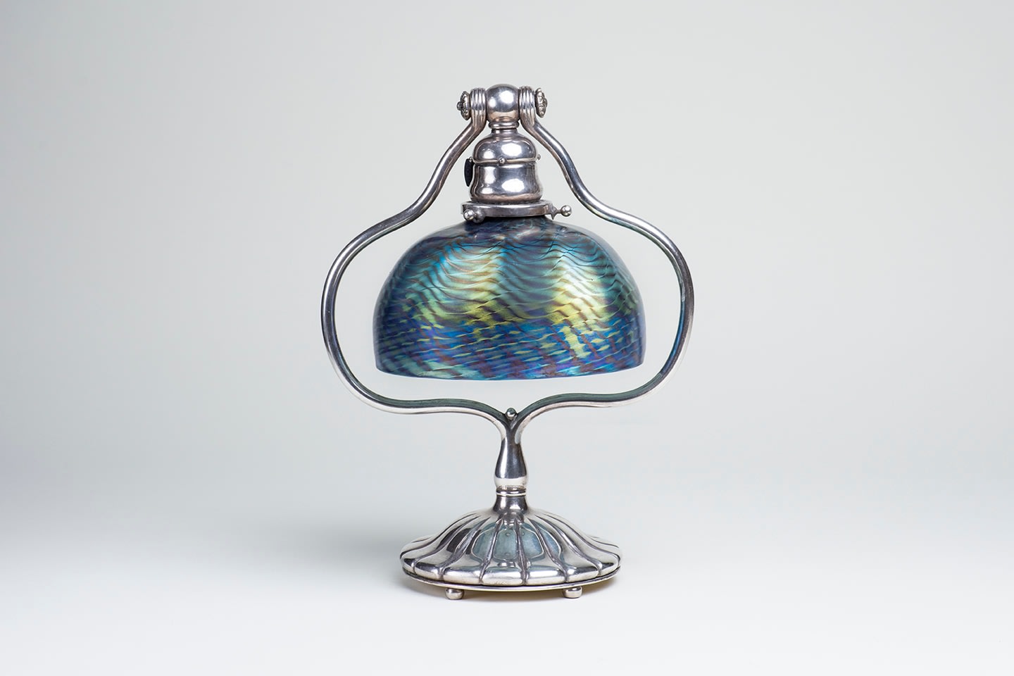 a tiffany studios desk lamp with a harp or bell style base in silvered bronze, holding a dome shaped shade of blue iridescent tiffany glass decorated all over with a silvery-gold wave motif. The shade is suspended from the harp base in such a way as to allow the angle of the shade and light to be adjusted.