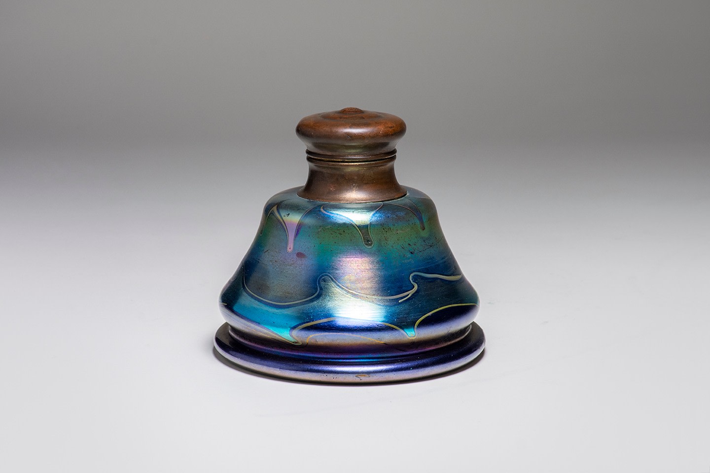 an inkwell formed by blown blue iridescent tiffany glass, with two bands of horizontal decoration of pulled hooked motifs in light blue, with overall silvery blue iridescence. The inkwell features a hinged bronze lid.