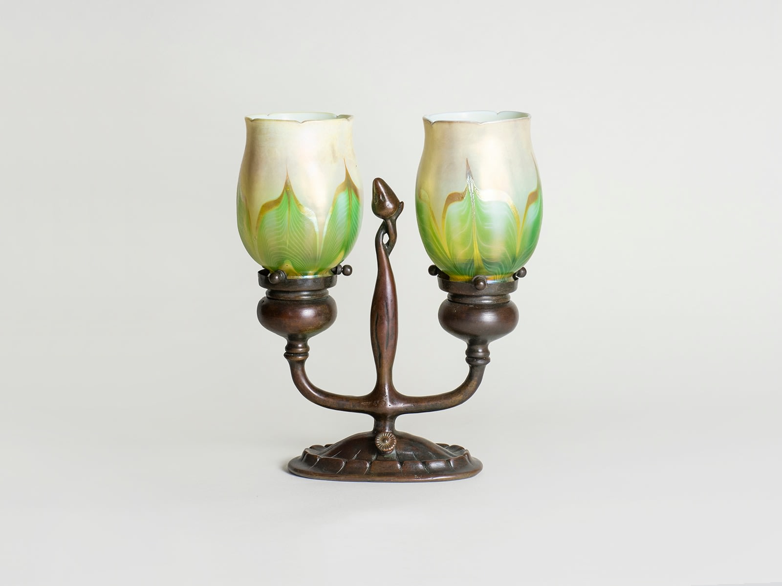 an electrified candle lamp with a bronze base, with a central handle in the form of a stylized bud, on either side framed by two tiffany favrile glass &quot;tulip&quot; shades in opalescent white glass with green and iridescent pulled feather motif