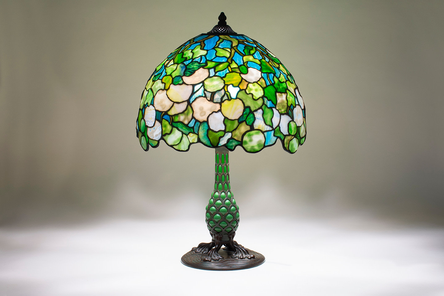 a leaded glass tiffany lamp, the shade depicting a snowball or viburnum bush with rounded blossoms formed by mottled white tiffany glass with green rippled glass leaves against a background representing blue sky, on a &quot;pineapple&quot; model lamp base with green tiffany glass blown through an openwork armature of metal.