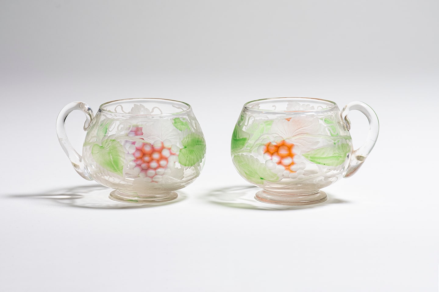 a pair of small rounded punch glasses formed by clear tiffany glass decorated with motif of pink champagne grapes and green grape leaves which have been wheel carved with additional decorative details