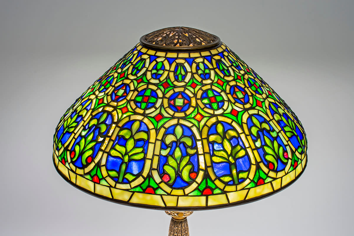 a detail of a conical leaded glass tiffany lamp shade in the &quot;venetian&quot; pattern, with alternating oval medallions depicting green palmettes and fleur de lys against blue background, with geometric motifs in red and green glass