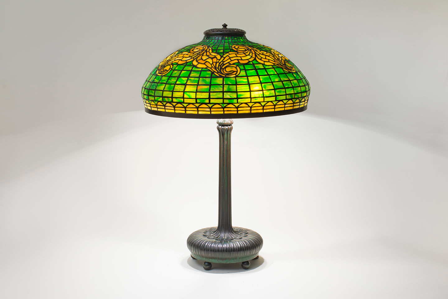 an original tiffany lamp with leaded glass shade with geometric background of gridded green glass with decoration of swag motif in contrasting yellow glass, on bronze lamp base with a round cushion-like foot