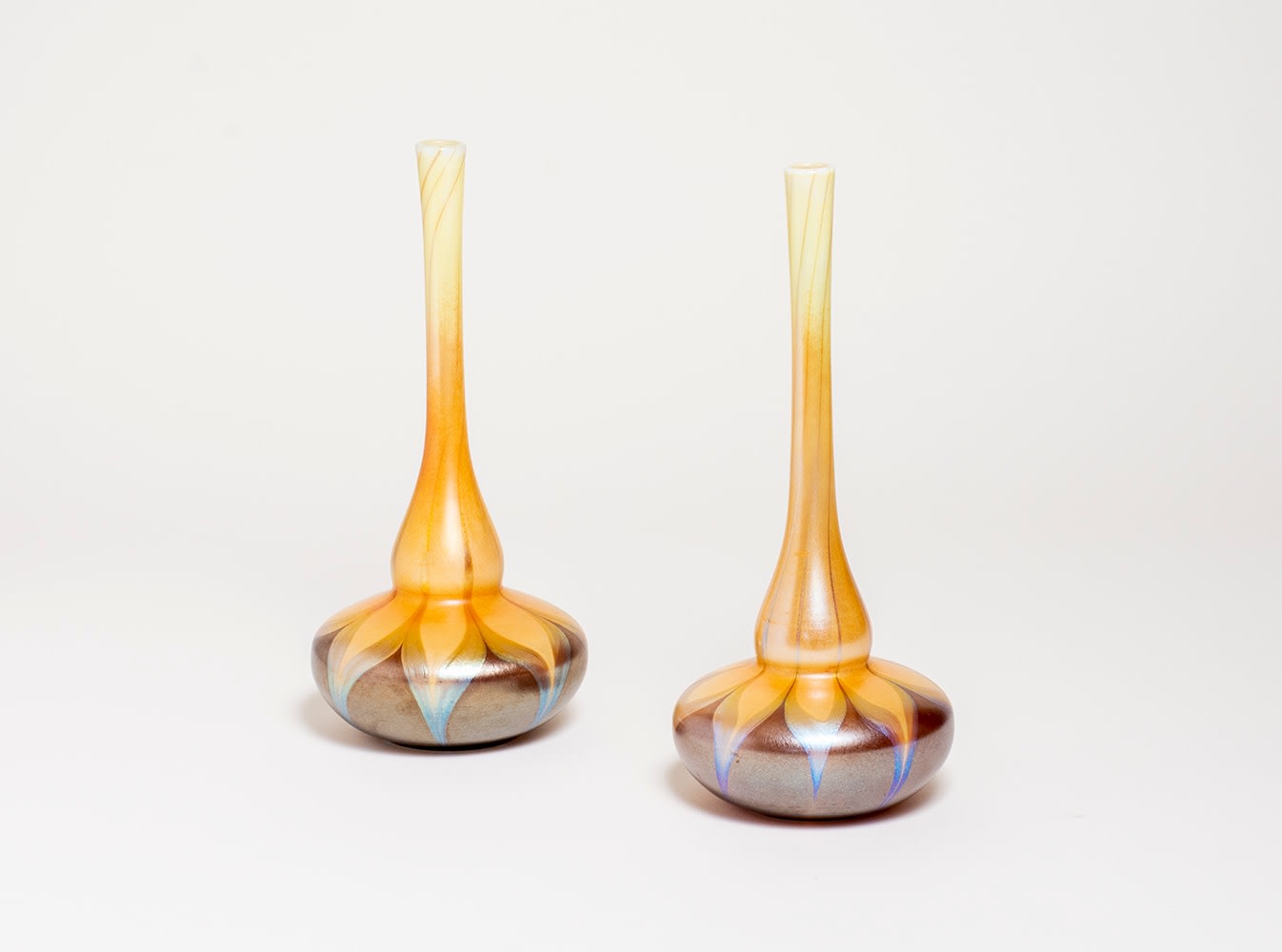 a pair of bottle neck tiffany favrile glass vases with very thin  necks and rounded bases, the base in a caramel brown glass, the neck in a pale yellow, divided by a band of swag decoration in iridescent silvery blue