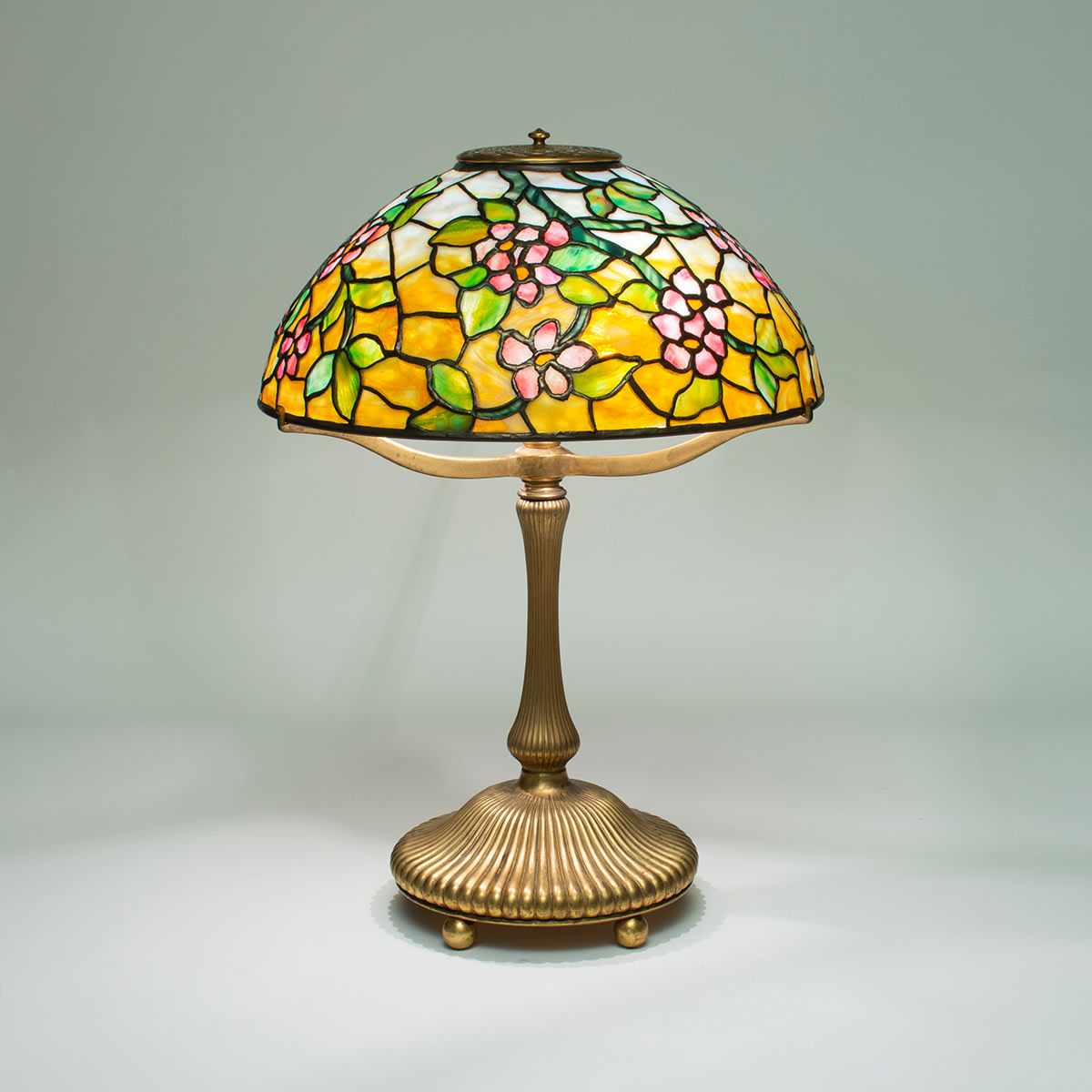 a tiffany lamp with leaded glass shade depicting greenish branches with pink mottled apple blossoms and green leaves against a background of mottled yellow glass, on a base of gilt bronze with vertical fluting