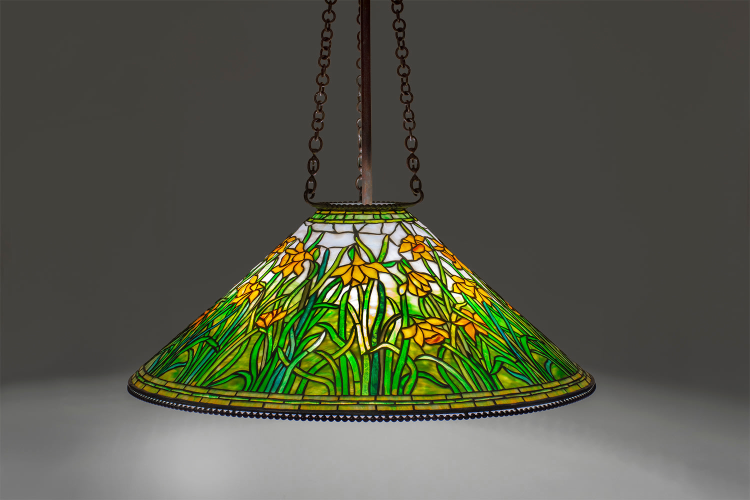 a cone shaped hanging tiffany lamp with beaded upper and lower rims, hanging from three chains, depicting in leaded glass a series of stylized yellow daffodils in mottled yellow glass, the lower edge of the shade filled with the daffodil's green spiky stems and leaves, the upper portion of the sky a milky opalescent white glass