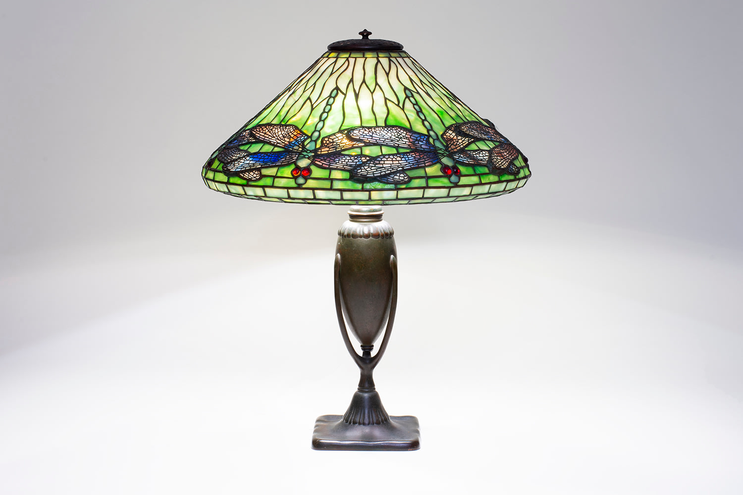 a leaded glass tiffany lamp depicting dragonflies with pale teal bodies, their wings formed by glass streaked with blue and purple with pierced metal filigree over top, against a background of soft green glass which is more saturated near the lower edge of the cone-shaped shade, on a bronze table lamp base