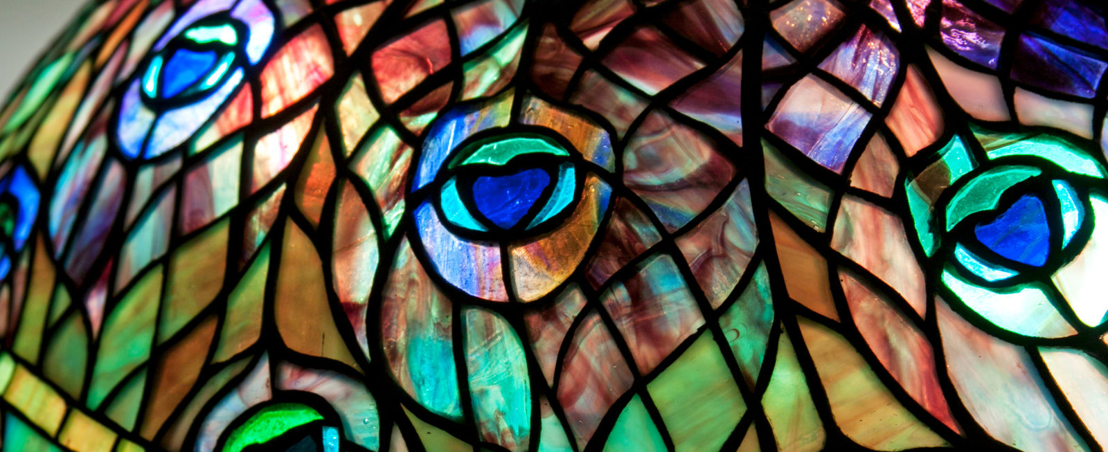 a leaded glass tiffany lamp, close up detail showing the striated coloration of the tiffany glass in shades of purple, teal, and golden amber, depicting the eyes of peacock feathers
