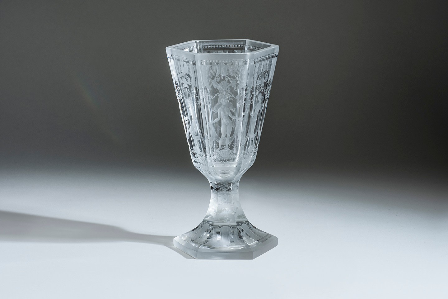 A footed chalice of clear glass, the six sided &quot;cup&quot; decorated on each side with a unique motif of a nude female of classical style with garlands and classical motifs, the stem and flared foot with engraved geometric decoration