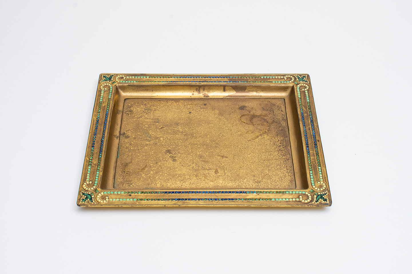 a shallow bronze rectangular tray in gold patina, the flattened raised rim with decoration in inset enamel ranging from yellow to blue, with stylized yellow fleur de is at each corner.