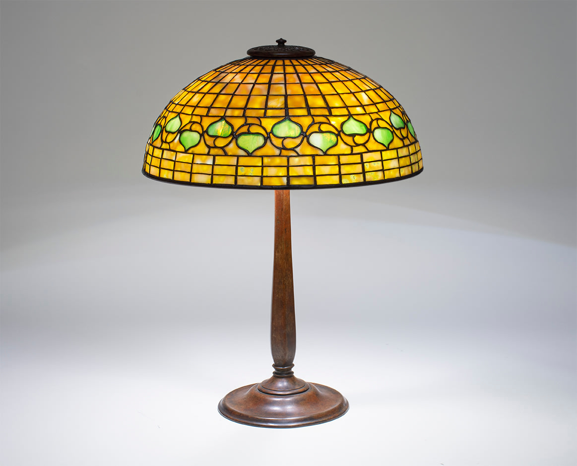 a geometric tiffany studios lamp, leaded glass shade in the &quot;acorn&quot; pattern, the background a pale creamy yellow grid of glass with a horizontal decorative band of stylized leaves alternating pointing up and down in green glass