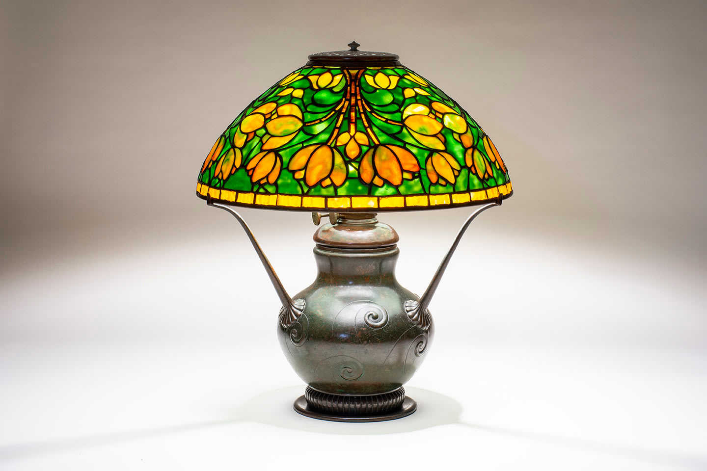 an early tiffany lamp with leaded glass shade depicting stylized yellow-orange tulips in symmetrical arcing pattern against a mottled green background, supported by a three-armed early tiffany lamp base meant for oil or kerosene with an urn-shaped body decorated with swirling motif