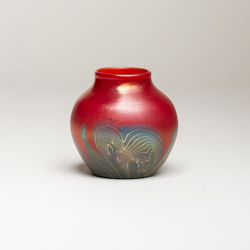 a favrile glass cabinet vase by tiffany studios, the small rounded body in a deep scarlet red glass with soft surface iridescence, the lower portion decorated with a gunmetal striated swirling abstract motif.