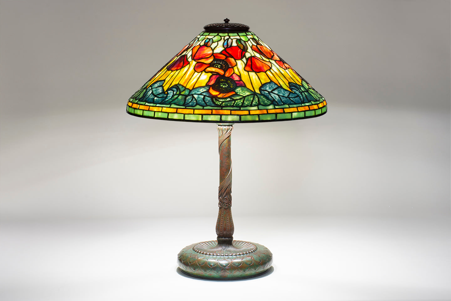 a leaded glass Tiffany Lamp depicting red poppies with pierced metal filigree mounted on top of the glass representing the centers of the flowers, against a background of variegated yellow glass, with poppy buds in blue-red glass near the top of the shade, the lower edge of the shade with green leaves, the vines formed by pierced metalwork mounted to the interior of the shade.