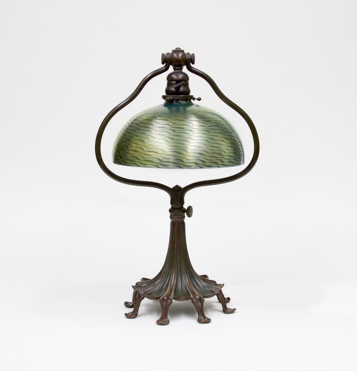 a tiffany desk lamp dating from around 1910, a footed bronze base with stylized leaf design with a bronze harp supporting a blown green tiffany glass damascene shade with iridescent silver wave decoration