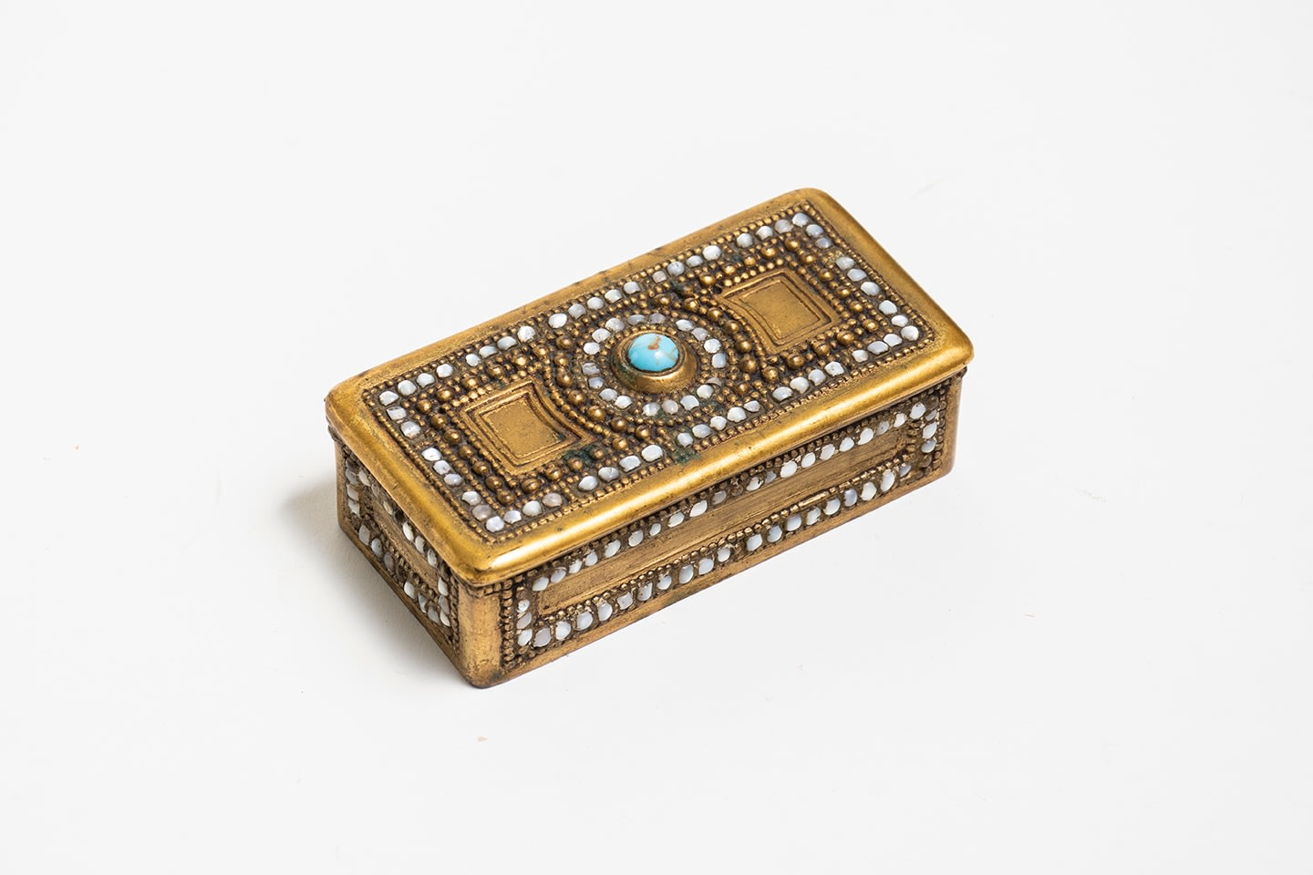 A small rectangular gilt bronze stamp box with raised beading on the flat lid, with small inset pieces of iridescent white mother of pearl and a glass bead at the center in teal glass with brown swirls meant to recreate a piece of turquoise.