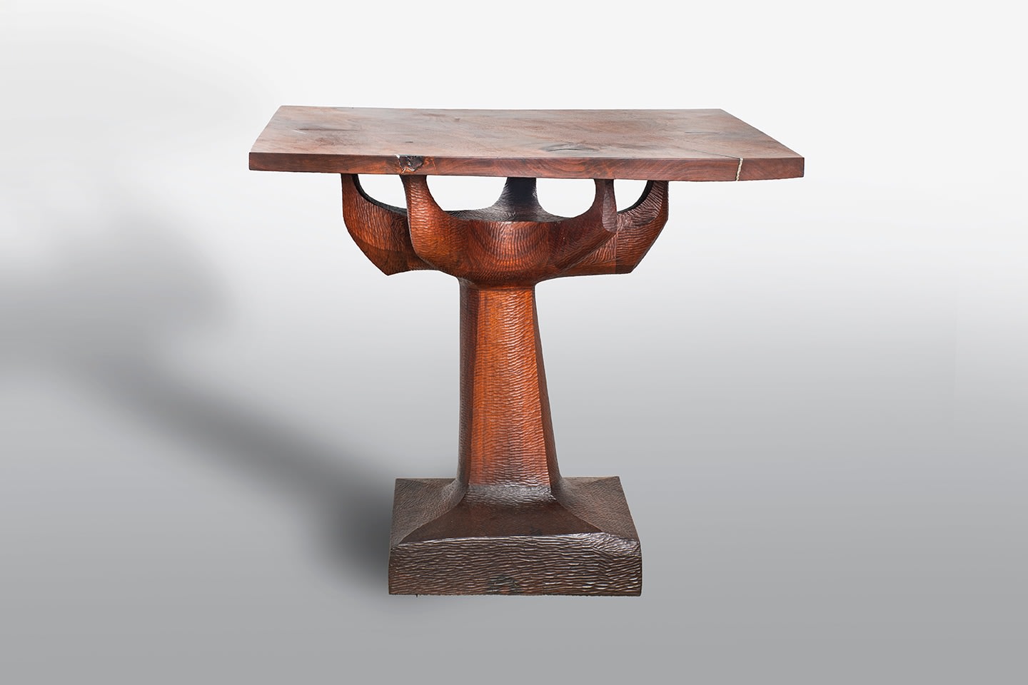 an early walnut table by Wendell Castle, the base formed by several thick blocks of wood which have been joined together and then chip carved to form a squared base from which a &quot;tree trunk&quot; styled stem rises and spreads to form five curved branches, which support a flat squared thick table top with areas of aluminum inset into the grain of the wood