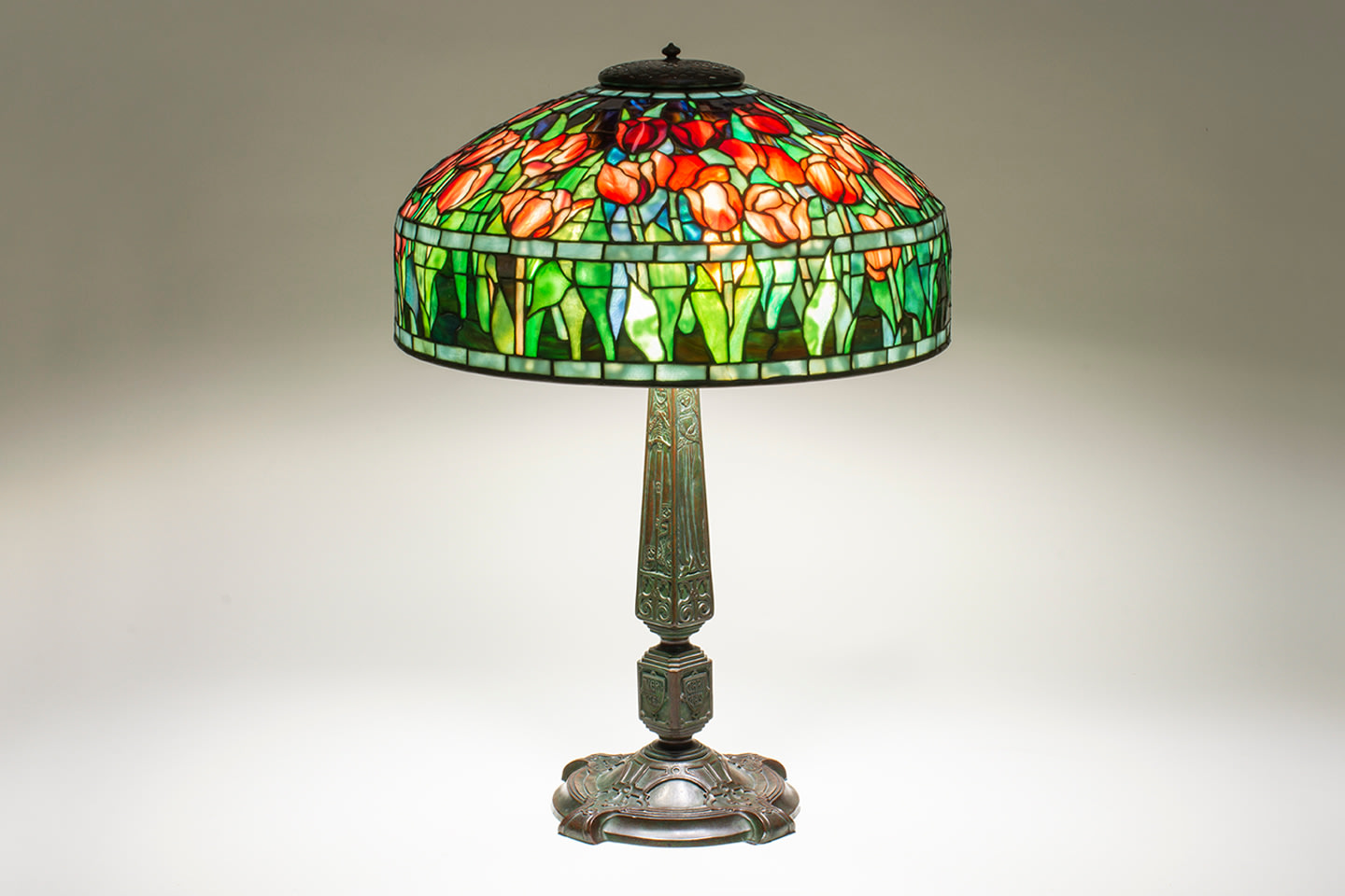 an original tiffany lamp featuring a domed leaded glass shade depicting a naturalistic field of red tulips with green leaves, with purple sky, on a bronze base decorated with stylized medieval depictions of the four virtues as maidens