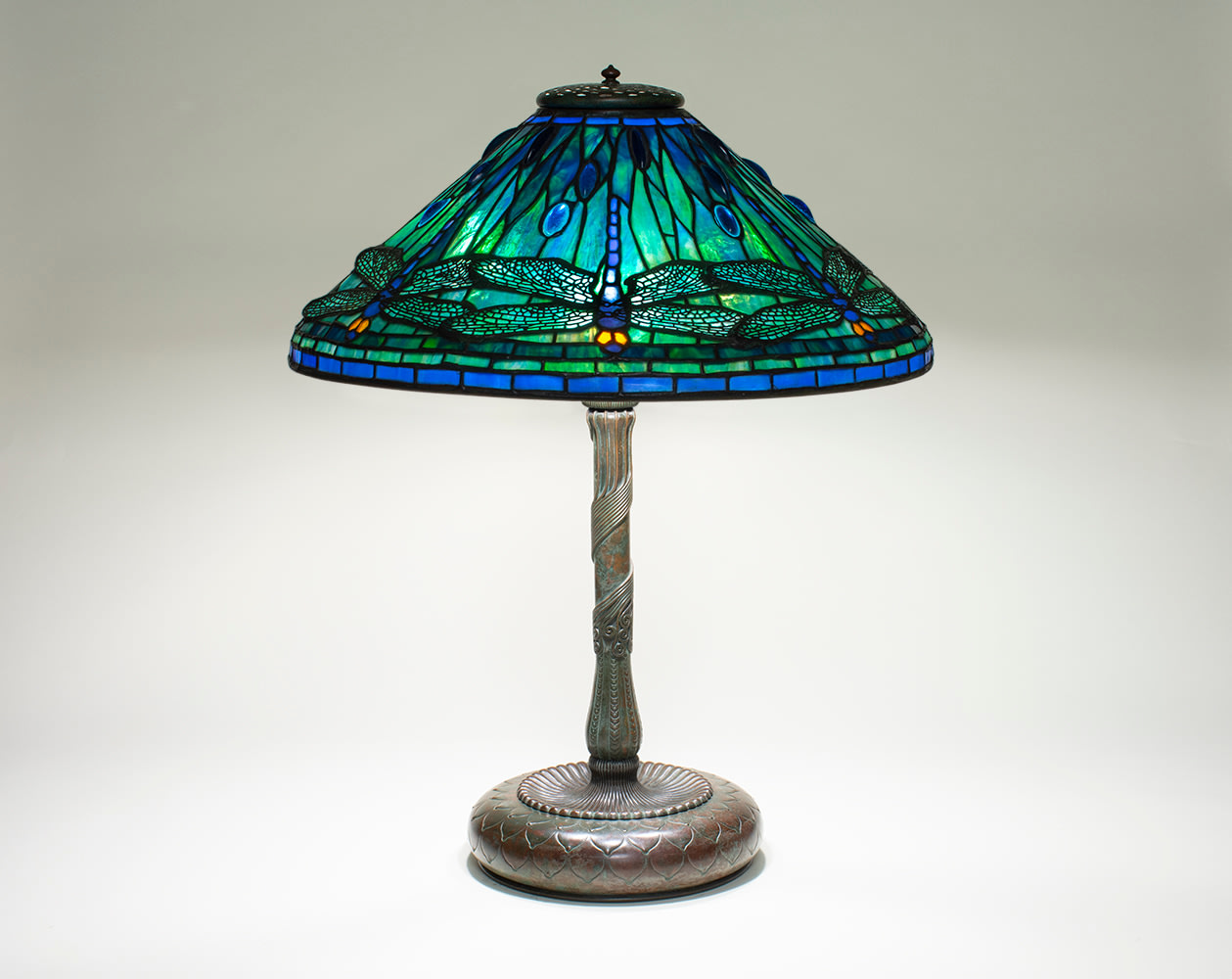 a leaded glass tiffany lamp depicting blue dragonflies with filigree wings and golden eyes, against a background of swirling blue-green glass representing water, on a bronze stick base with wirework decoration