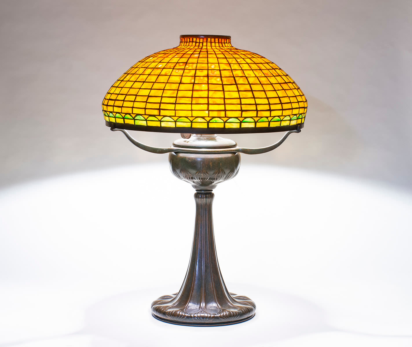 a geometric tiffany lamp dating from 1904, on a tyler bronze base in dark brown patina, with three thin bronze arms supporting a leaded glass shade formed by orange mottled tiffany glass in a grid, the lower border with semi-circular green glass border.