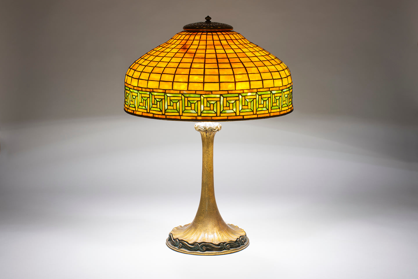 a tiffany lamp with gilt bronze base of fluted form with scrolling detail at the foot, supporting a shade formed by gridded orange-amber mottled tiffany glass with a decorative horizontal border at the lower edge depicting a greek key pattern in green and cream glass